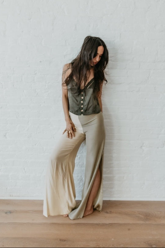 Woman in a green top and beige pants standing against a white brick wall.