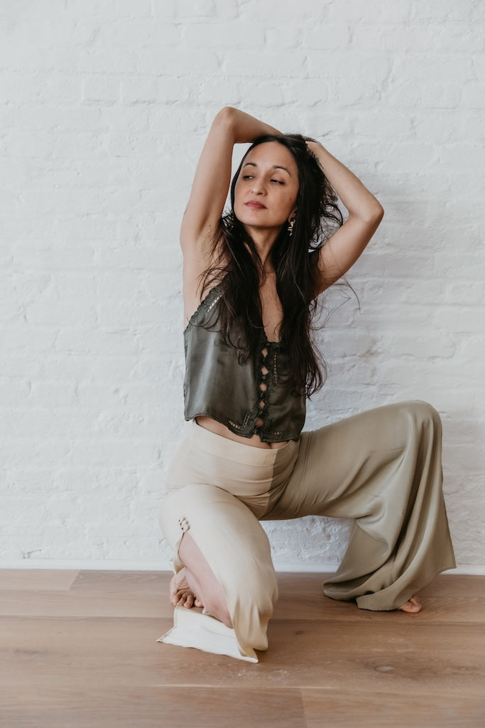 Woman wearing licorice embroidered top posing with hands above head against a white brick wall.