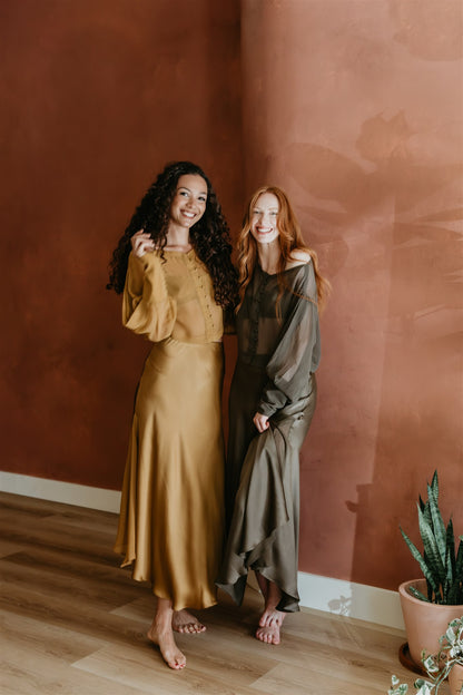 Two women in elegant dresses standing against a warm-toned wall with a plant in the corner.