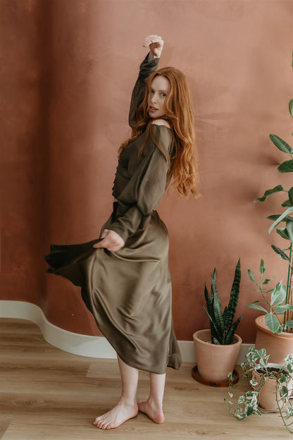 Woman in a long, dark green dress standing against a brown wall with potted plants on a wooden floor.