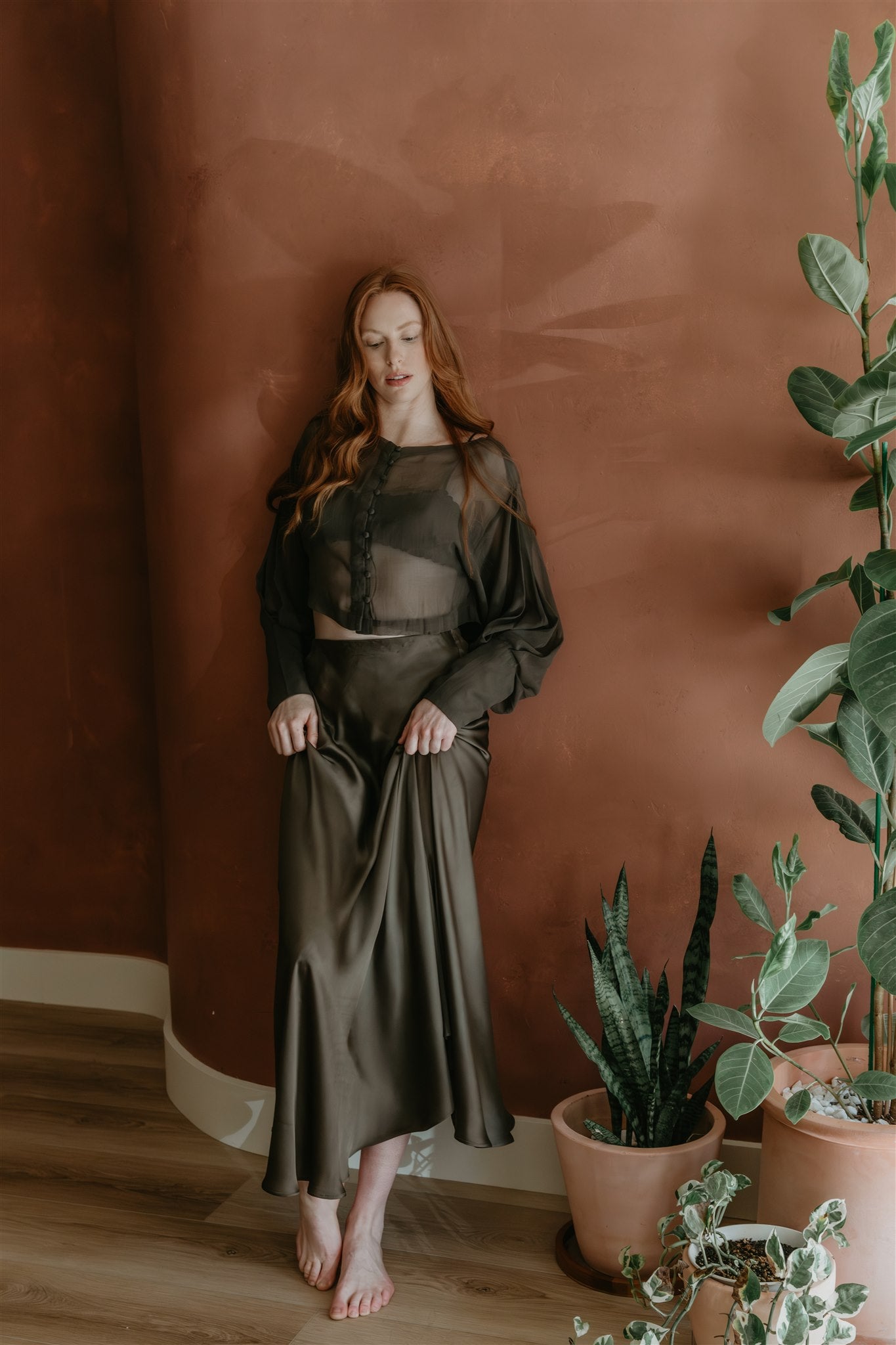 Woman in a dark green skirt standing in a room with plants against a brown wall.