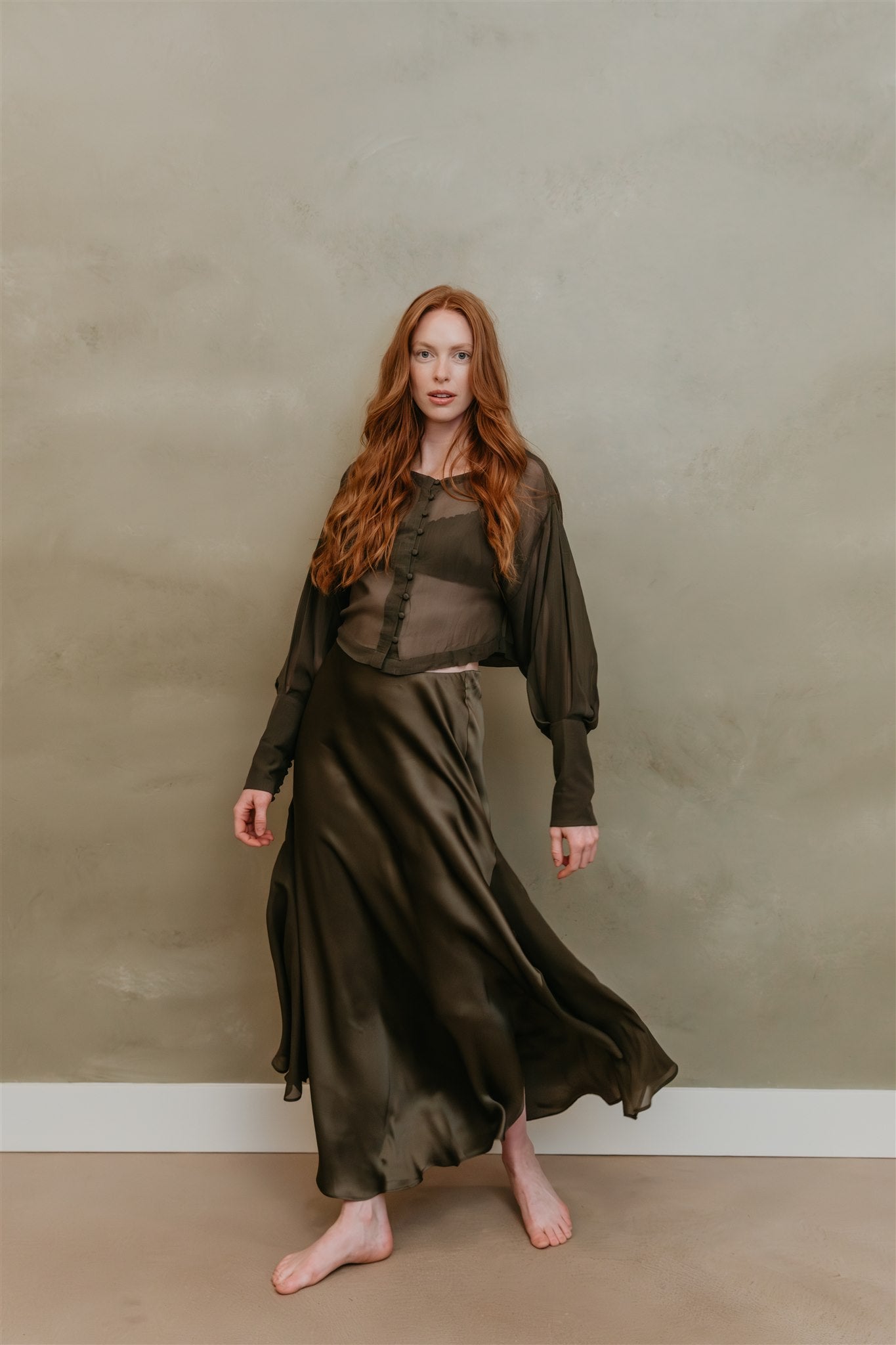 Woman wearing a long, dark green skirt against a plain wall.
