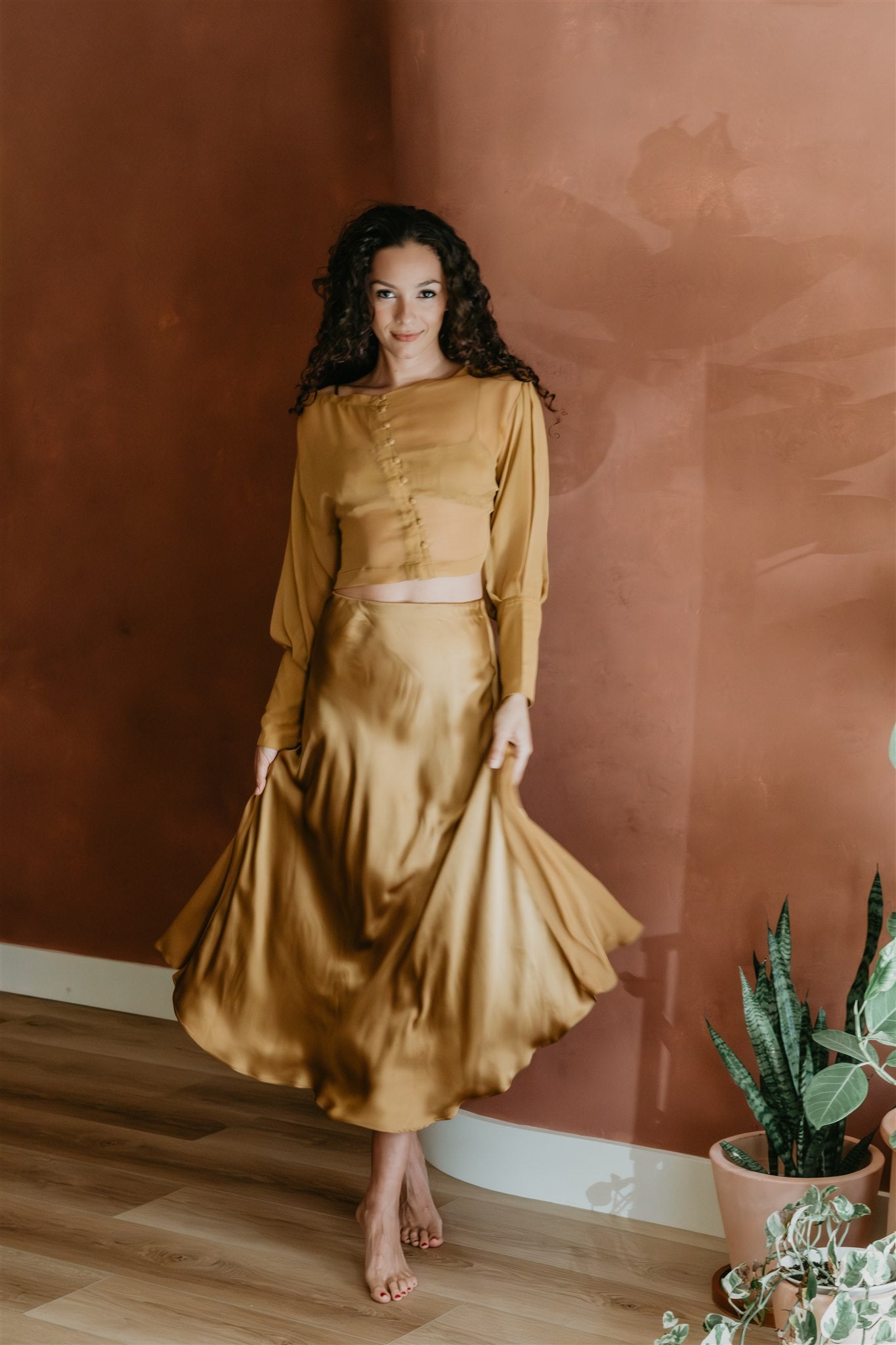 Woman in a mustard gold outfit standing against a brown wall with plants around.