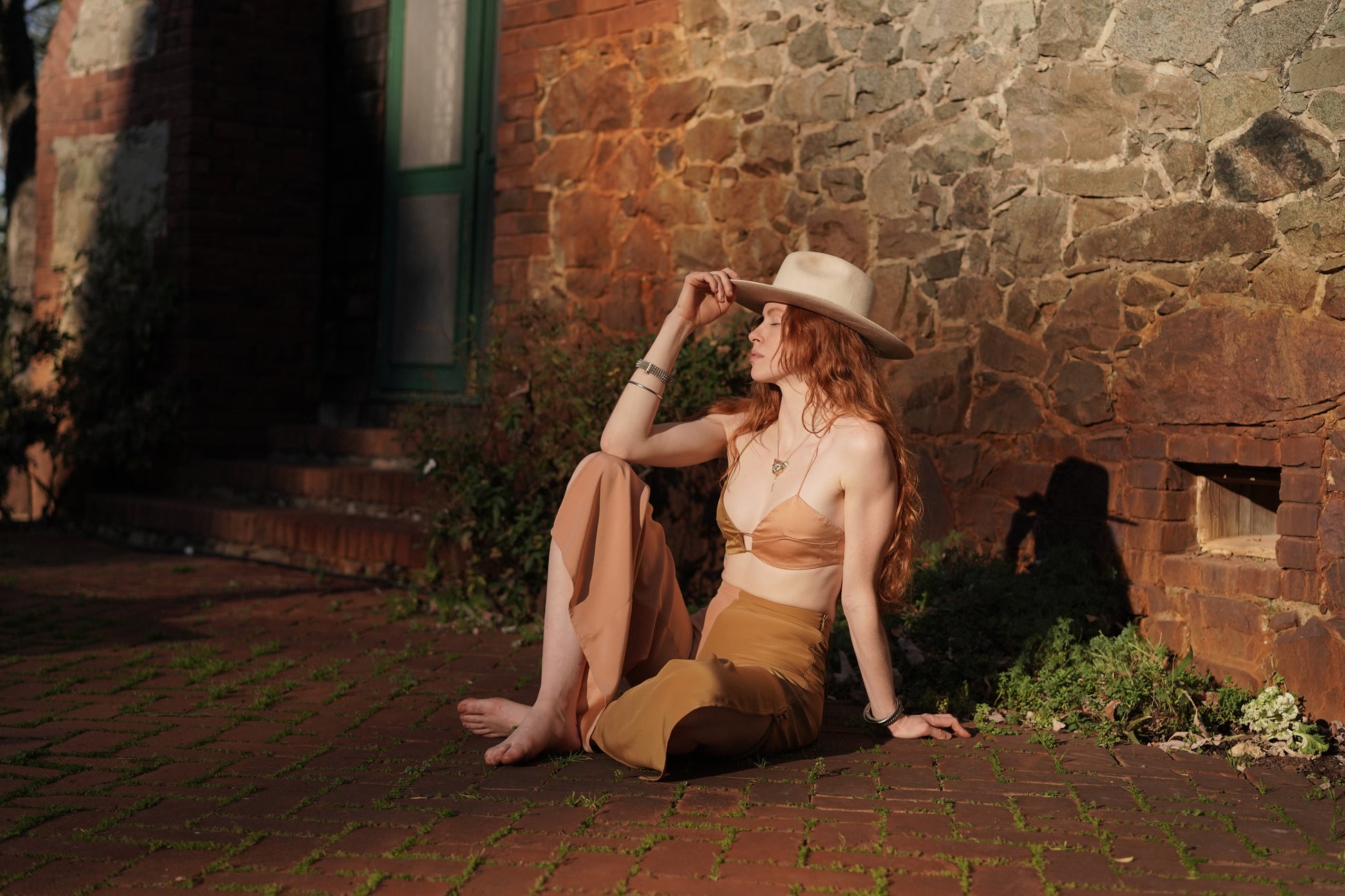 Woman sitting on a brick path in front of a stone wall wearing a hat and peach color silk pants.