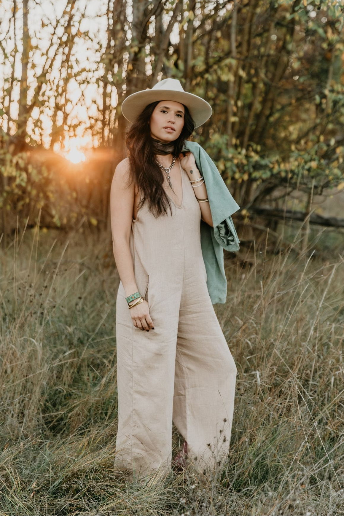 Woman in a beige linen jumpsuit and hat standing in a field with sunset in the background