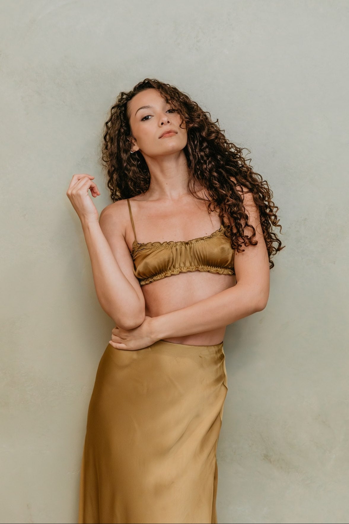 Woman wearing a gold satin Bralette and skirt against a plain background