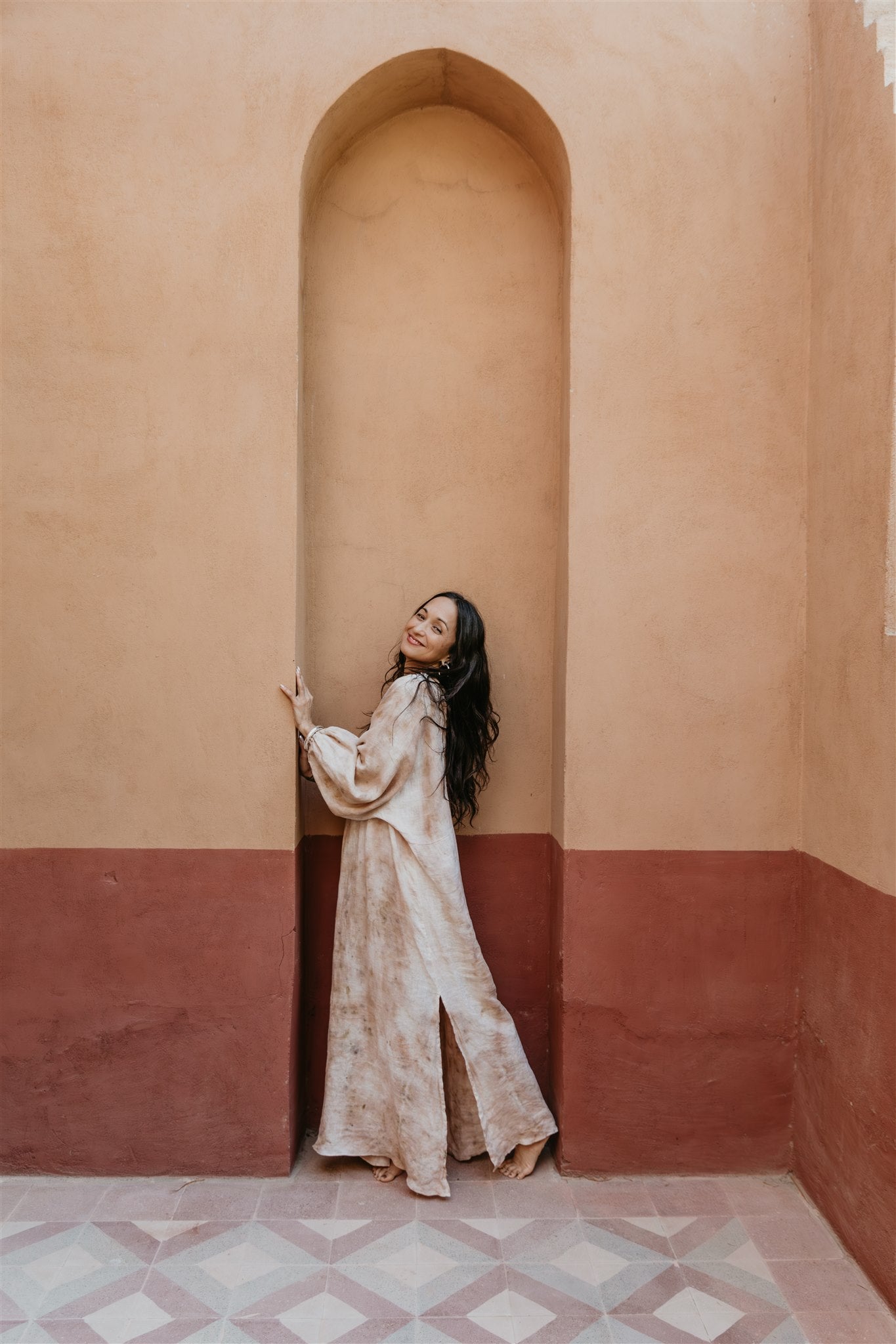 Woman in a long, light-colored dress standing in a room with a pink wall and geometric floor.