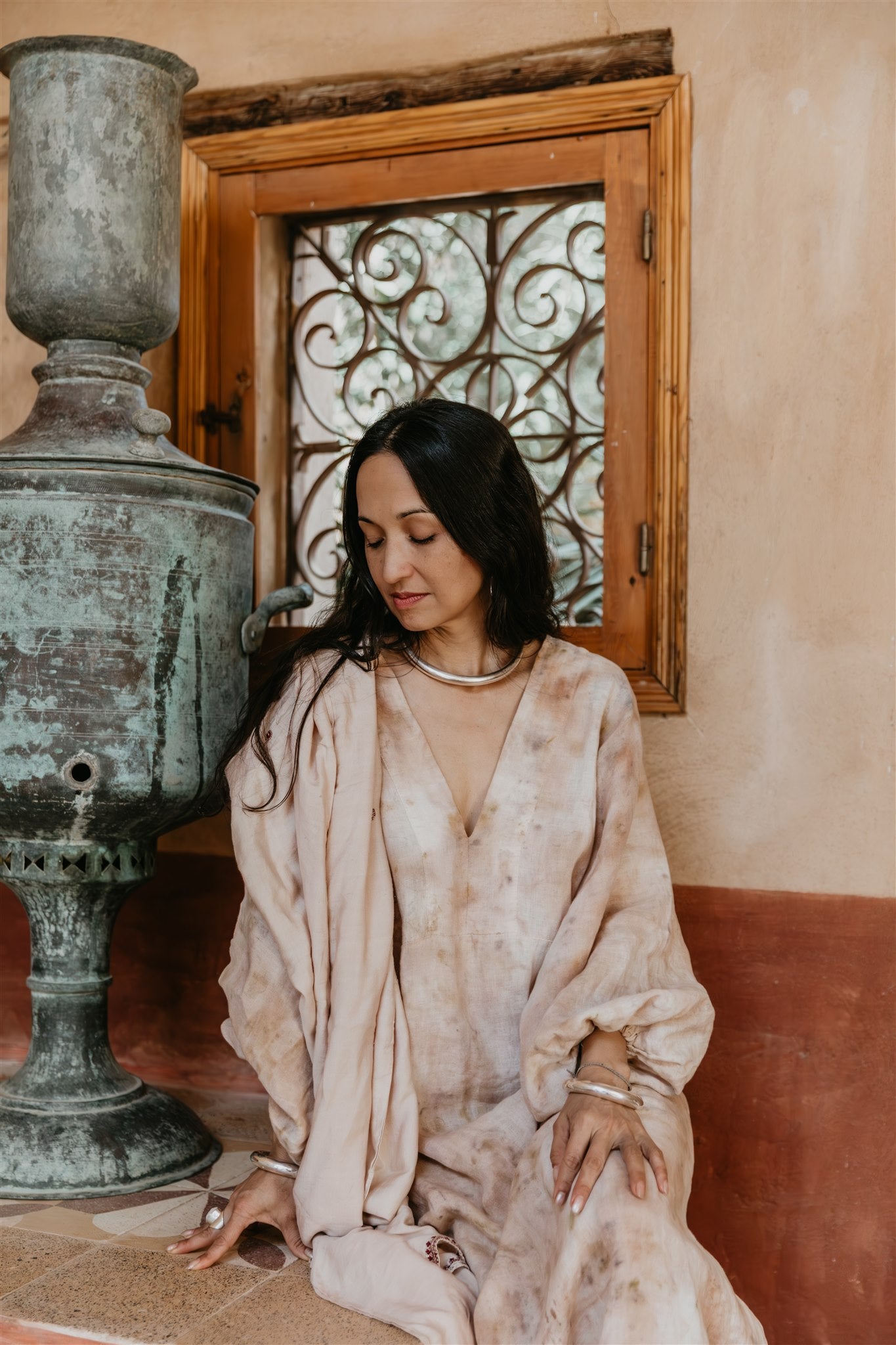 Woman wearing Nomad Linen kaftan by Payet Maison with deep V neckline and ballon sleeves, sitting next to a large vase in a room with decorative elements.