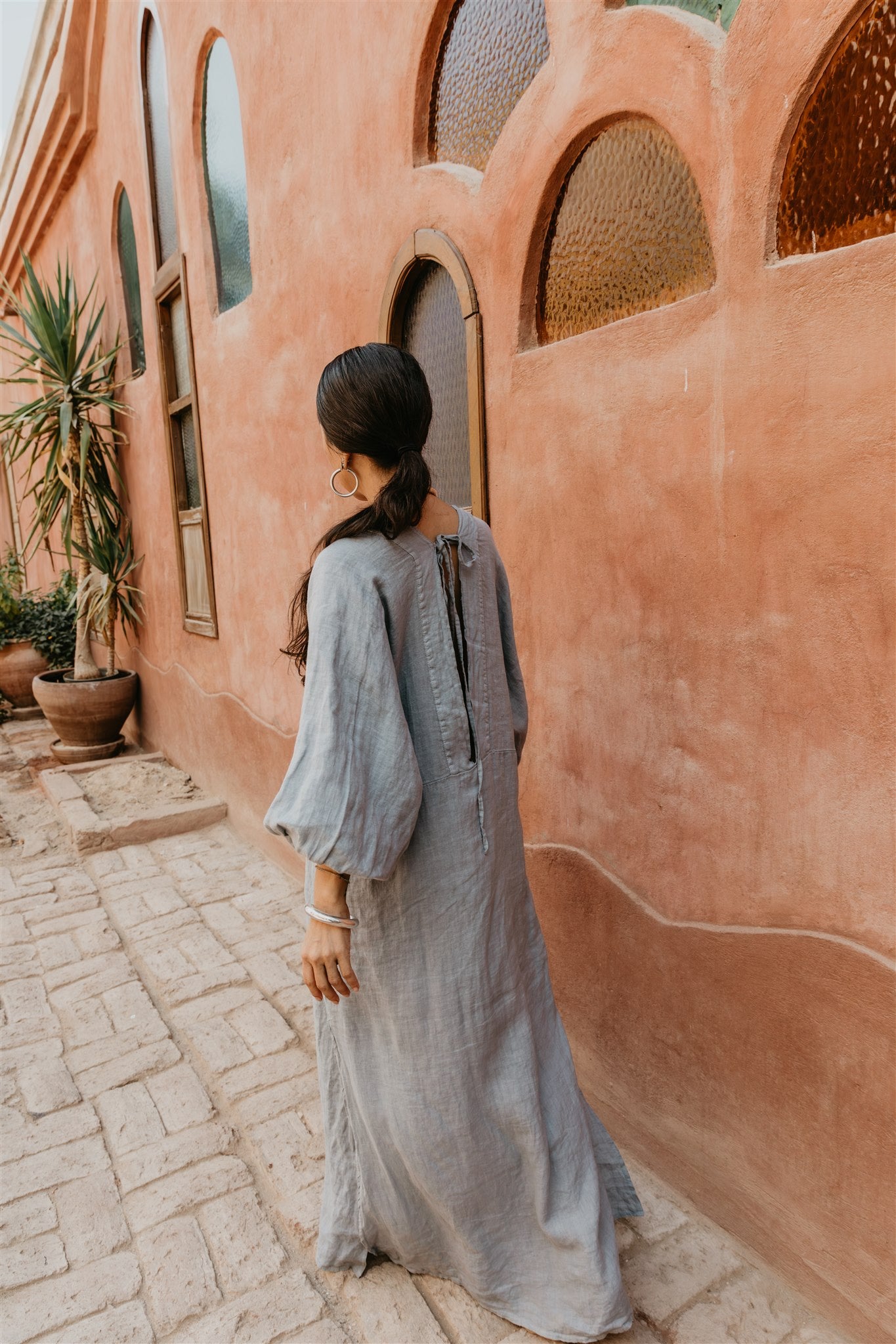 Woman in a long light blue dress standing in front of a textured pink wall with arched windows.