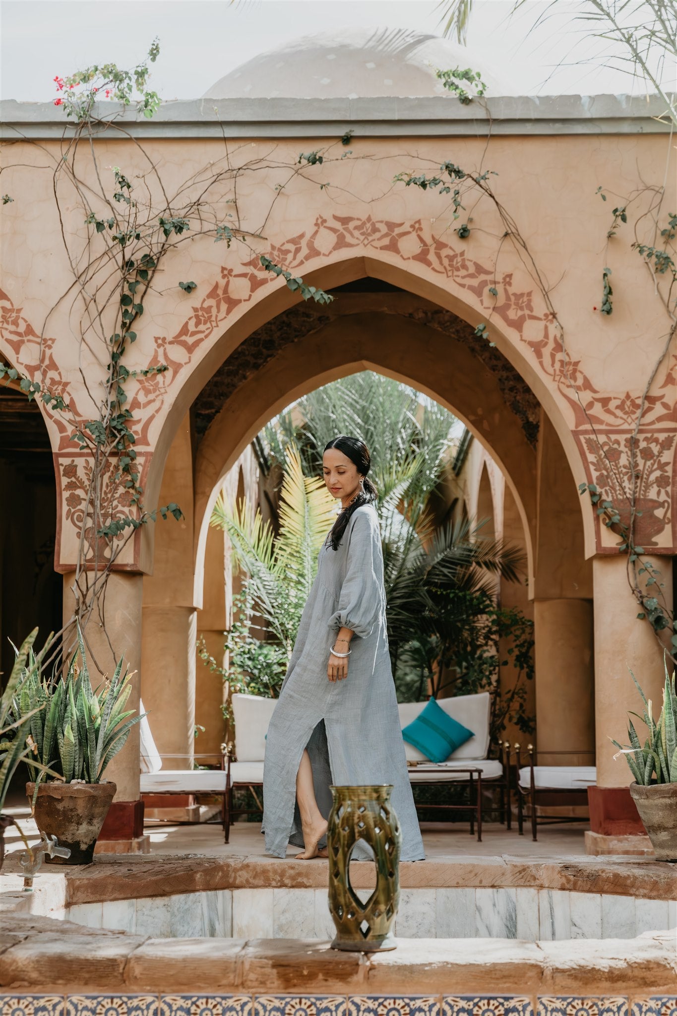 Woman in a linen  long dress standing in a decorative archway with plants and furniture.
