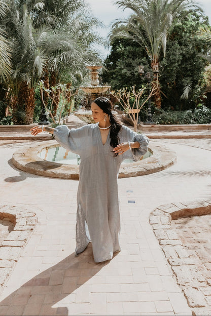 Woman in a light gray dress standing in a garden with palm trees and a fountain.