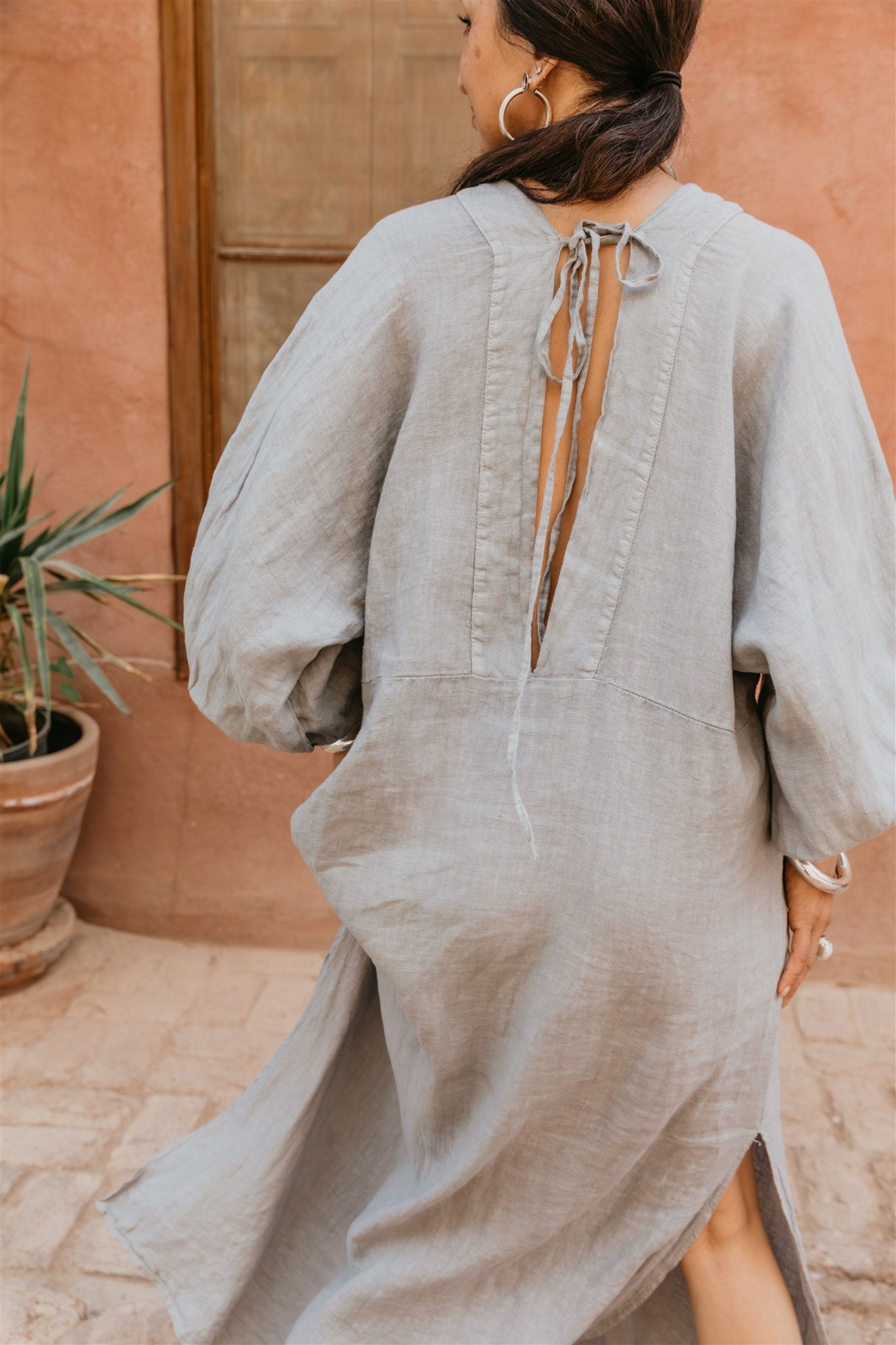 Woman wearing a light gray dress with a back cutout, standing against a terracotta wall.
