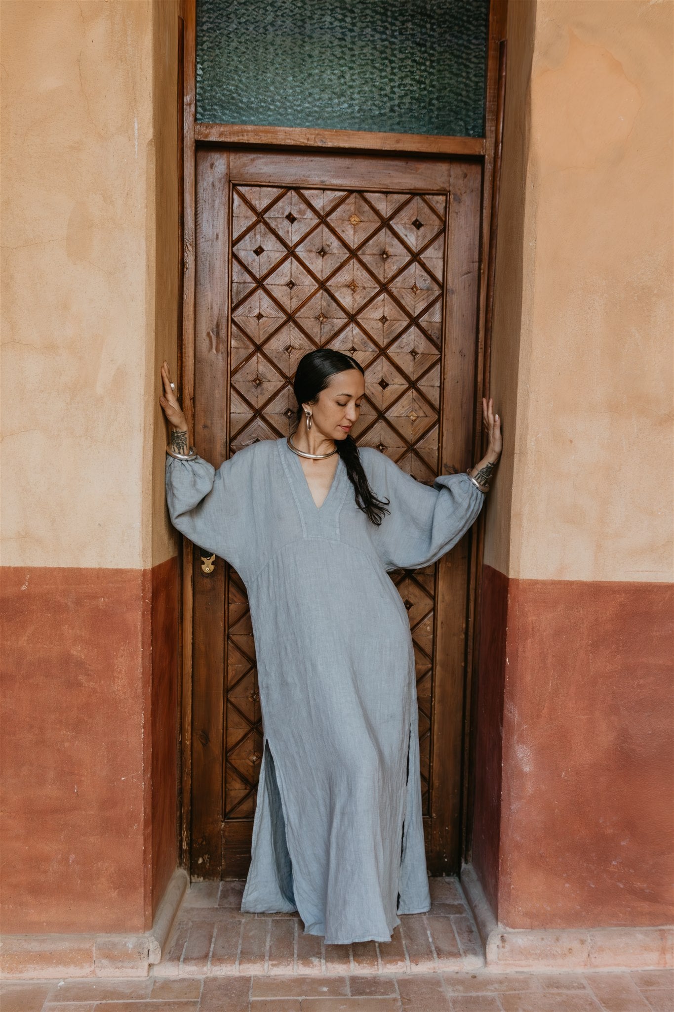 Woman in a long, light blue dress standing in a doorway with a patterned wooden door.