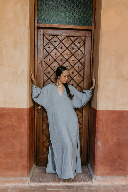 Woman in a long, light blue dress standing in a doorway with a patterned wooden door.