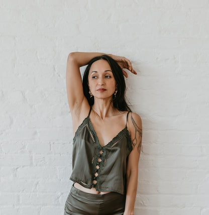 Woman wearing a green silk satin top with embrodery leaning against a white brick wall
