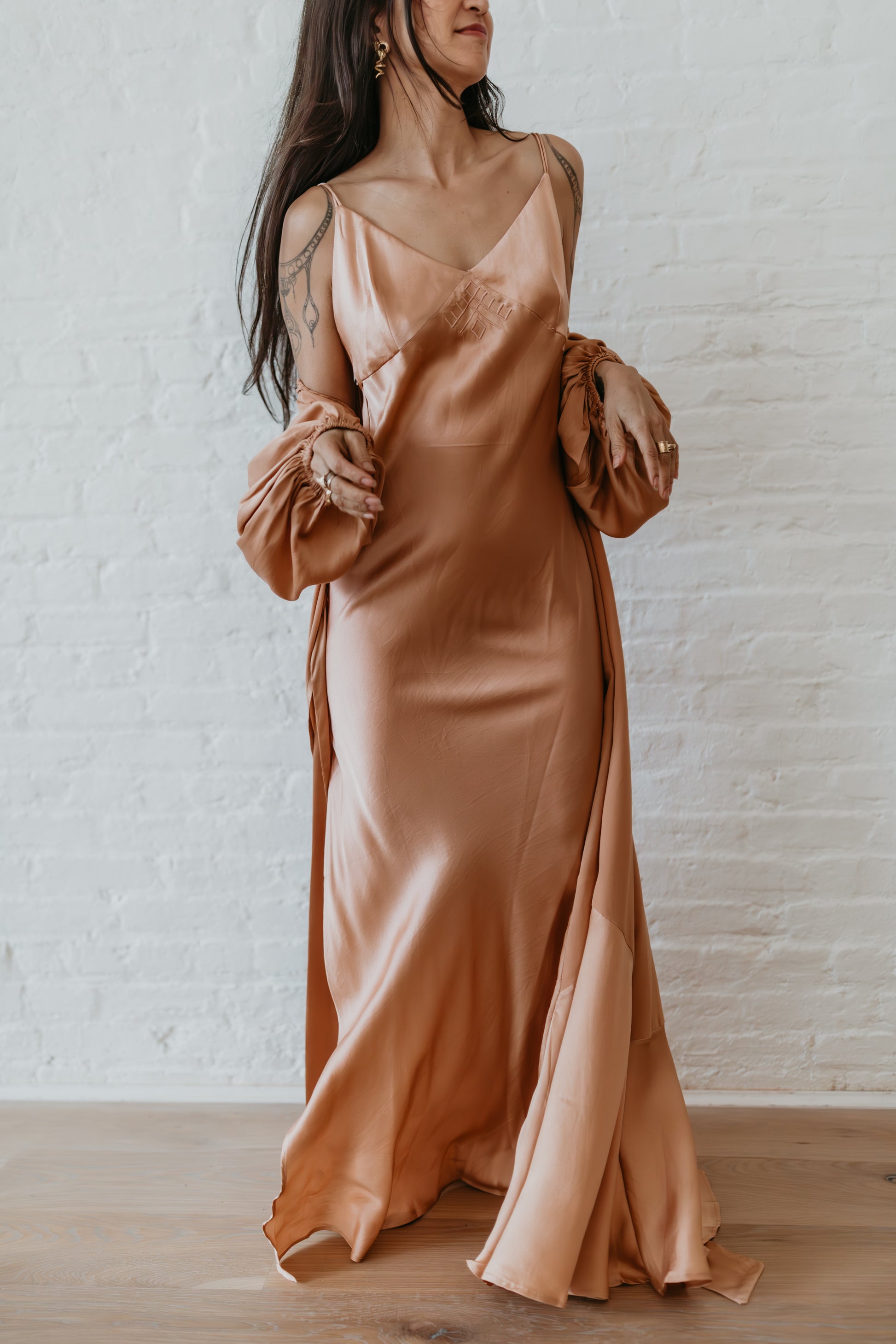 Woman wearing a long, flowing dress with ruffled sleeves against a white brick wall.