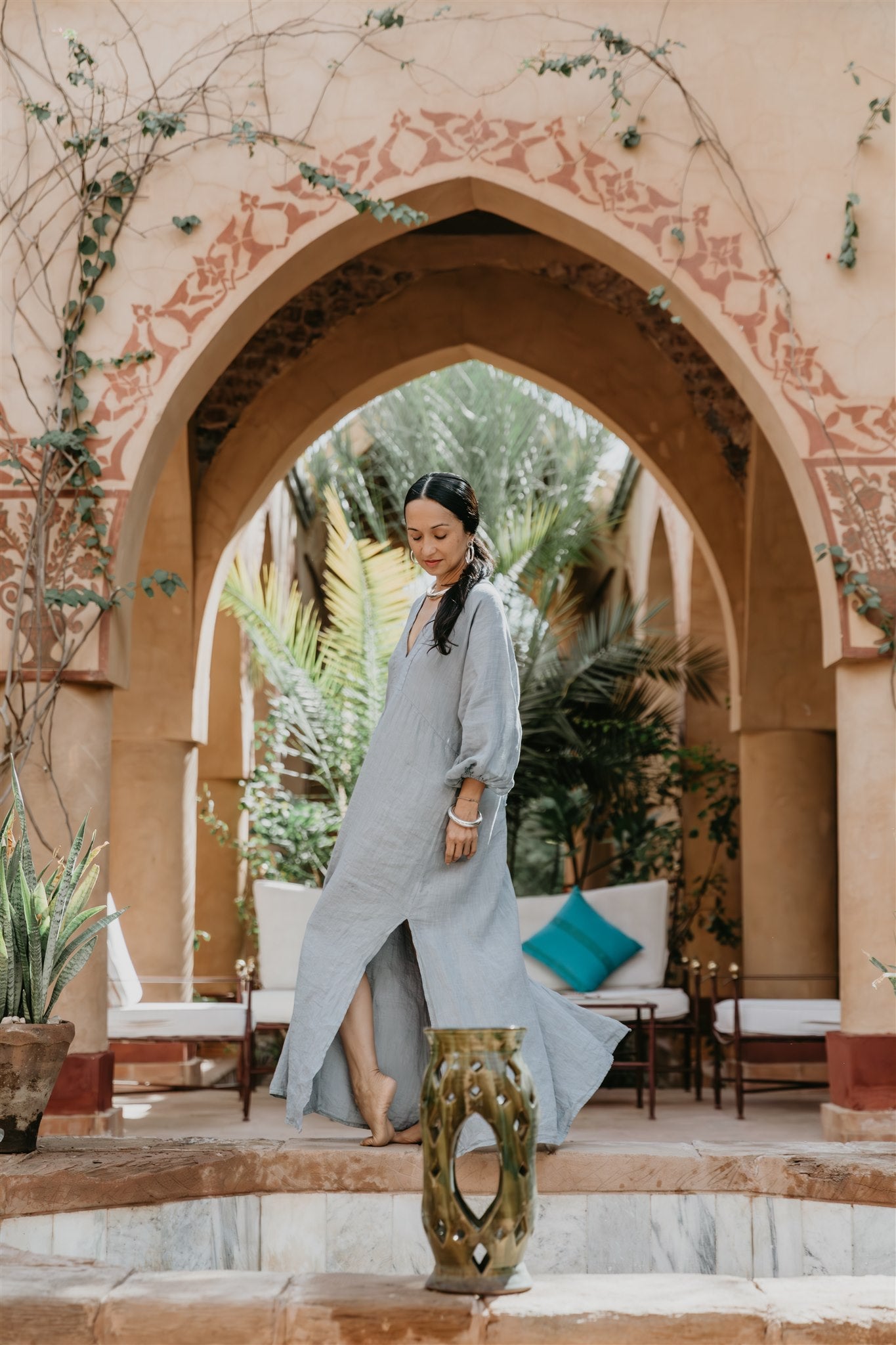 Woman in a long blue dress standing under an ornate archway in a garden setting.