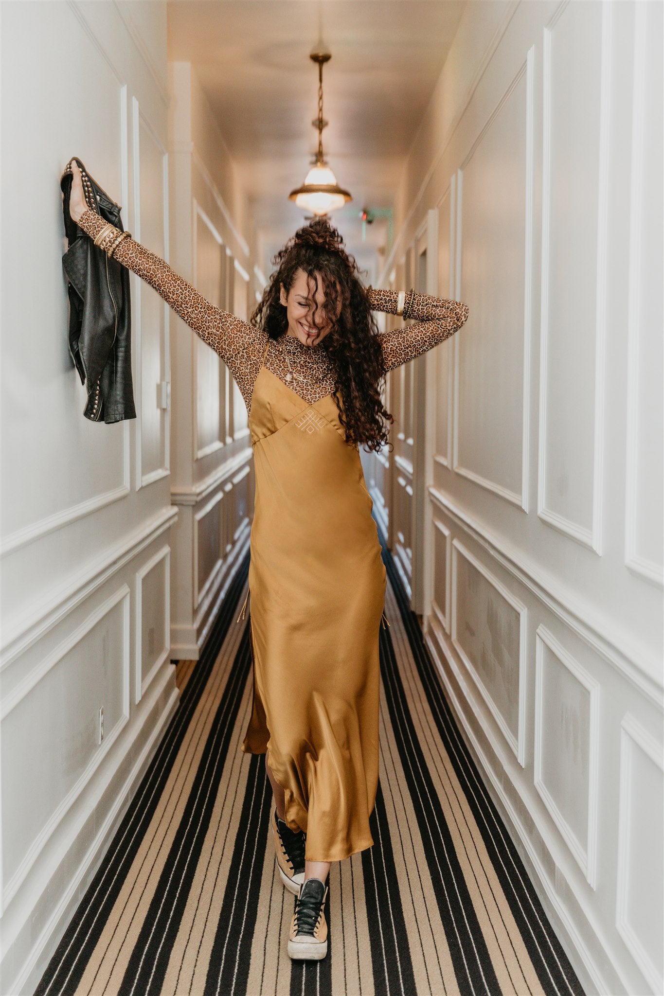 Woman in a mustard yellow silk dress standing in a hallway with decorative walls and striped floor.