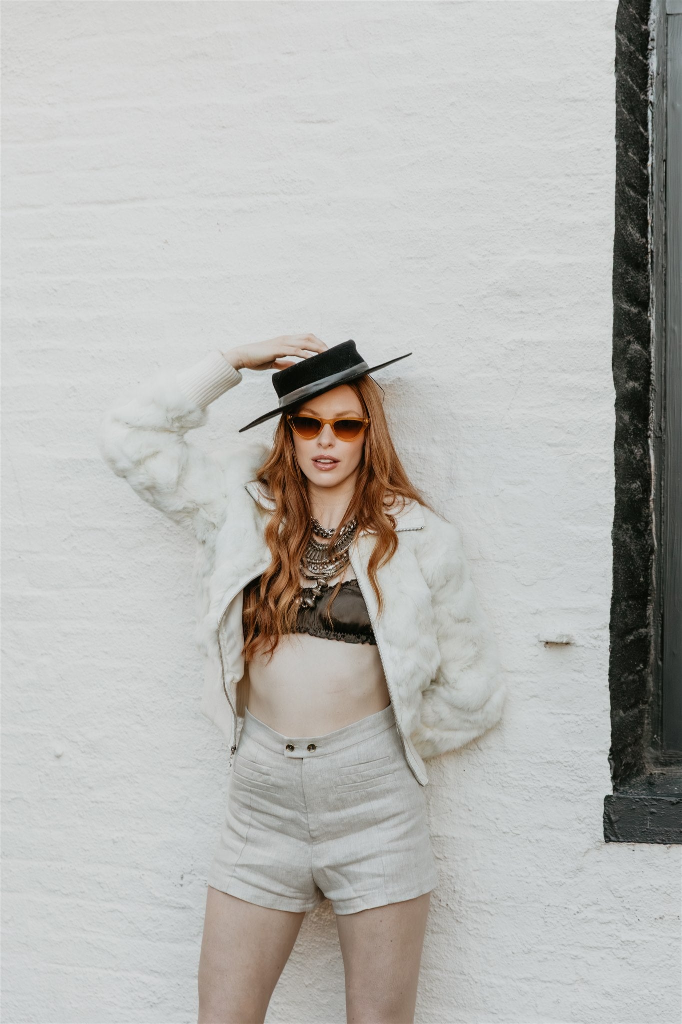 Woman wearing a white fur coat, black hat, sunglasses, and beige shorts against a white brick wall.