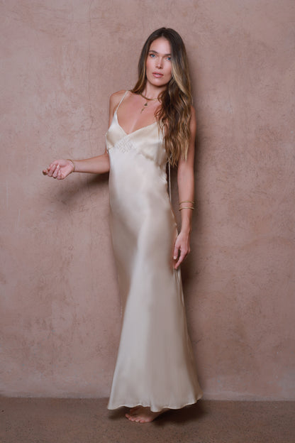 Woman wearing a long, light-colored satin silk dress against a beige wall.